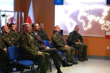 Grupa żołnierzy w polskich mundurach wojskowych i mężczyzna w garniturze siedzą na niebieskich krzesłach podczas prezentacji z mapą świata na ekranie.