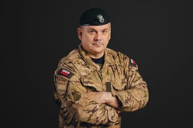 Mężczyzna w polskim mundurze wojskowym z beretem stoi z założonymi rękami na ciemnym tle.