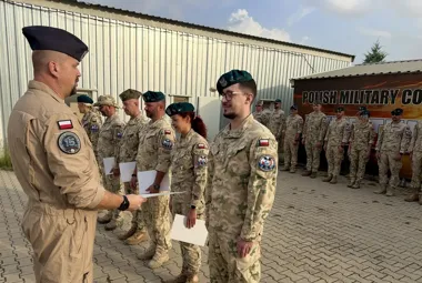 Żołnierze w mundurach stoją w szeregu przed budynkiem z napisem „POLISH MILITARY CO” podczas uroczystego wręczania dokumentów przez oficera.