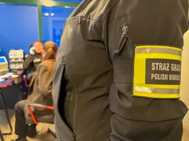 Funkcjonariusz Straży Granicznej w pomieszczeniu służbowym, w tle inni ludzie przy biurku.