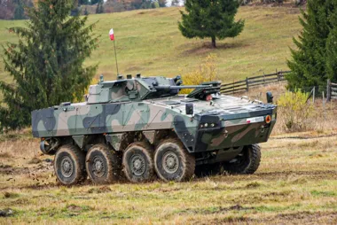 Wojskowy kołowy transporter opancerzony z polską flagą porusza się po polnej drodze w pagórkowatym terenie.