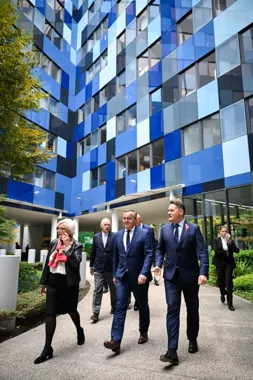 Grupa osób w formalnym stroju spaceruje przed nowoczesnym budynkiem o niebiesko-białej elewacji.