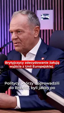 Mężczyzna w granatowym garniturze siedzi przy stole z mikrofonem i książką, w tle fioletowo-czarne pionowe pasy.
