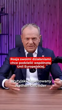 Mężczyzna w garniturze siedzi przy drewnianym stole z mikrofonem, w tle fioletowa ściana z pionowych listew.