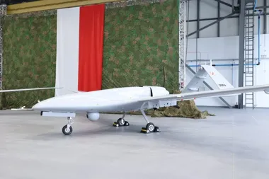 Biały bezzałogowy aparat latający stoi na posadzce w hali, za nim zielona siatka maskująca i flaga w kolorach biało-czerwonych.