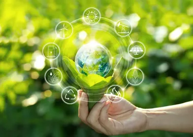 Dłoń trzyma zielonego liścia z kulą ziemską, otoczoną ikonami odnawialnych źródeł energii, na tle rozmytej zieleni.