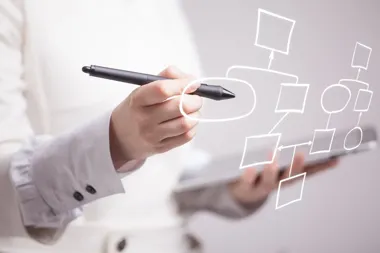 Osoba w białej koszuli rysuje długopisem schemat blokowy na przezroczystej powierzchni, trzymając tablet.