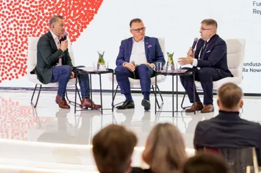 Trzech mężczyzn w garniturach uczestniczy w dyskusji panelowej na scenie przed publicznością.