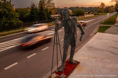 Pomnik geodety z teodolitem stoi przy krawędzi wieczornej drogi, po której przejeżdżają samochody.