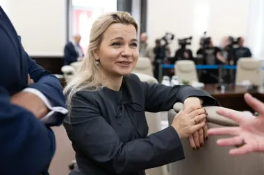 Kobieta w czarnej bluzce siedzi na krześle w sali konferencyjnej i rozmawia z osobami wokół niej.