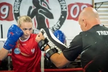 Bokserka w czerwonej koszulce i niebieskich rękawicach trenuje uderzenia z trenerem trzymającym łapy w klubie bokserskim.