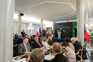 Uczestnicy konferencji siedzą przy stołach w sali z kolumnami, podczas gdy mówca występuje przy pulpicie na tle ekranu z napisami.