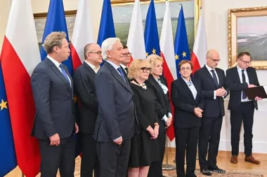 Grupa osób w formalnym stroju stoi w sali reprezentacyjnej przed flagami Polski i Unii Europejskiej podczas uroczystości.
