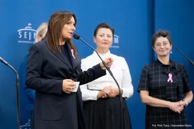 Kobieta w ciemnym garniturze przemawia do mikrofonu podczas konferencji prasowej w Sejmie, za nią stoją trzy inne kobiety z różowymi wstążkami.