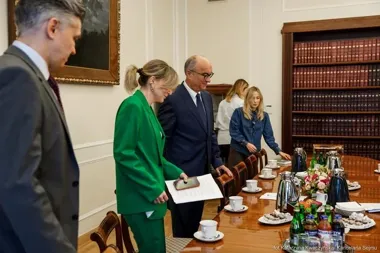 Grupa osób stoi przy nakrytym stole w eleganckim pomieszczeniu z biblioteczką, jedna z kobiet trzyma dokumenty i telefon.