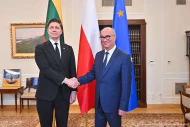 Dwaj mężczyźni w garniturach witają się uściskiem dłoni w reprezentacyjnym pomieszczeniu przed flagami Litwy, Polski i Unii Europejskiej.