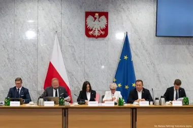 Sześć osób siedzi przy stole konferencyjnym w sali z polską i unijną flagą oraz godłem Polski na ścianie.