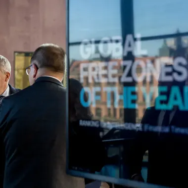 Dwaj mężczyźni w garniturach stoją przy szybie z napisem „Global Free Zones of the Year” podczas wydarzenia biznesowego.
