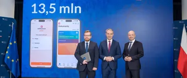 Trzej mężczyźni w garniturach stoją przed niebieską ścianką promującą aplikację mObywatel z napisem „13,5 mln” i grafikami telefonów, po bokach flagi UE i Polsk