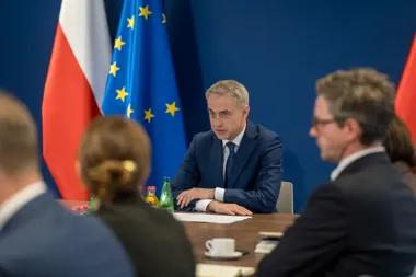 Mężczyzna w garniturze siedzi przy stole podczas spotkania z flagami Polski i Unii Europejskiej w tle.