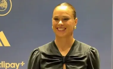 Uśmiechnięta kobieta w czarnej bluzce z kokardą pozuje podczas wydarzenia z logo sponsorów w tle.
