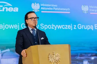 Mężczyzna w garniturze przemawia z drewnianego pulpitu z orłem podczas konferencji prasowej z logotypami Enea i ministerstw na tle.