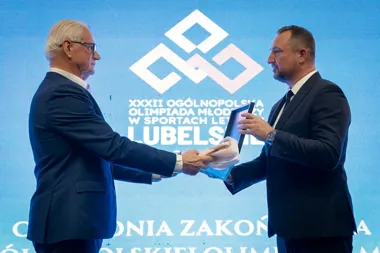 Dwaj mężczyźni w garniturach stoją na scenie przed niebieskim ekranem z logotypem XXXII Ogólnopolskiej Olimpiady Młodzieży w Sportach Letnich Lublin 2024 i wręc