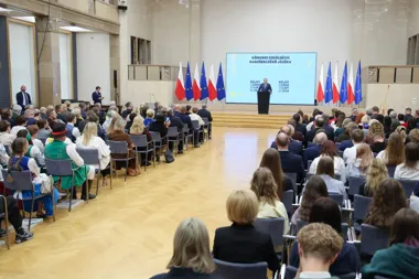 W auli zgromadzeni uczestnicy słuchają mówcy przy pulpicie podczas Kongresu Szkół Kaszubskiego Języka, za nim ekran z napisem w języku kaszubskim oraz flagi Pol