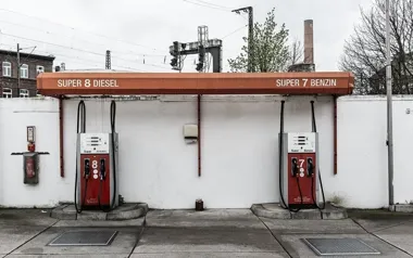 Stara stacja paliw z dwoma dystrybutorami oferującymi diesel i benzynę stoi przy białym murze pod pomarańczowym daszkiem.