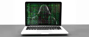 Laptop na szarym blacie, na ekranie widoczny sylwetka osoby w kapturze na tle zielonego kodu przypominającego efekt z filmu Matrix.