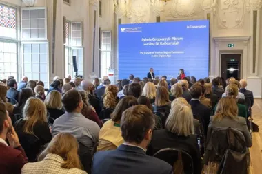 W reprezentacyjnej sali odbywa się spotkanie z udziałem publiczności, podczas którego dwie osoby prowadzą dyskusję na temat przyszłości ochrony praw człowieka w