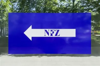 Błękitna tablica informacyjna z białym lewoskrętnym arrow i napisem NFZ stoi na chodniku przed zielonymi drzewami.