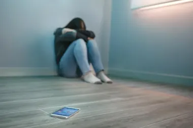 Osoba siedzi skulona w kąciu pustego pomieszczenia, a na podłodze leży smartfon.