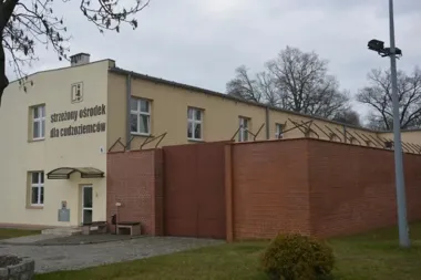 Budynek strzeżonego ośrodka dla cudzoziemców otoczonego ceglanym murem z drutem kolczastym.
