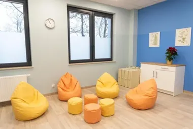 Pokój z kolorowymi pufami, oknami, zegarem i niską szafką, urządzony jako przestrzeń wypoczynkowa lub rekreacyjna.