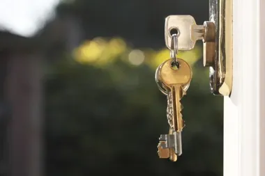 Klucze wiszą w zamku drzwi z otwartym wejściem, w tle rozmyty ogród z zielenią i żółtymi kwiatami.