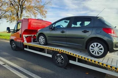 Szara osobowa hatchback stoi na platformie czerwonej lawety na asfaltowej drodze w otoczeniu pól i drzew.