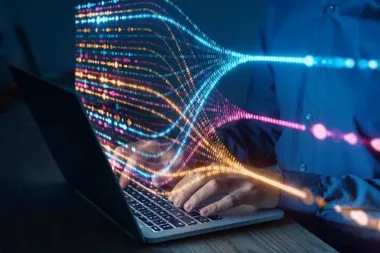 Osoba w niebieskiej koszuli pisze na laptopie, a z ekranu wypływają kolorowe, świetlne strumienie kodu binarnego.