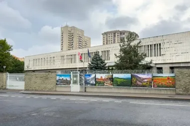 Budynek ambasady za kamiennym ogrodzeniem z plakatami krajobrazowymi, przed nim ulica i flagi.