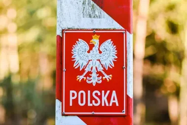 Czerwono-biały słup graniczny z tabliczką z orłem i napisem „Polska” na tle lasu.