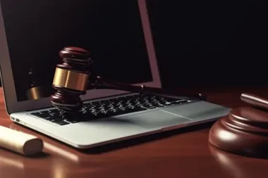 Młotek sędziowski leży na klawiaturze laptopa na drewnianym biurku, obok stoi jego podstawa.