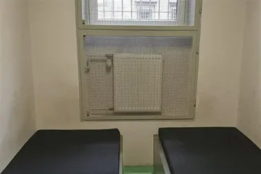 Wnętrze pustej, niewielkiej celi z dwoma czarnymi leżankami i oknem zabezpieczonym metalową siatką.