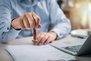Mężczyzna w niebieskiej koszuli przy biurku z laptopem odbija drewnianą pieczęcią dokument na clipboardie.
