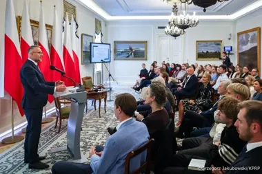 Mężczyzna w garniturze przemawia z mównicy do publiczności zgromadzonej w reprezentacyjnym salonie z polskimi flagami.