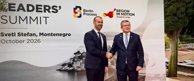 Dwaj mężczyźni w garniturach witają się uściskiem dłoni przed banerem zapowiadającym szczyt liderów w Czarnogórze w październiku 2026 roku.