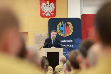 Mężczyzna w garniturze przemawia z mównicy do słuchaczy w mundurach w auli Akademii Pożarniczej pod polskim herbem.