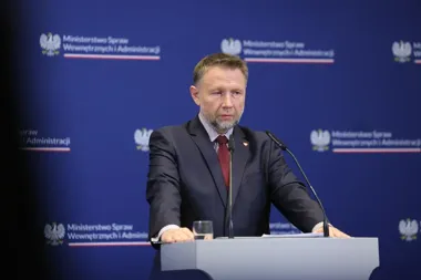 Mężczyzna w garniturze stoi za mównicą z mikrofonami podczas konferencji prasowej w Ministerstwie Spraw Wewnętrznych i Administracji.