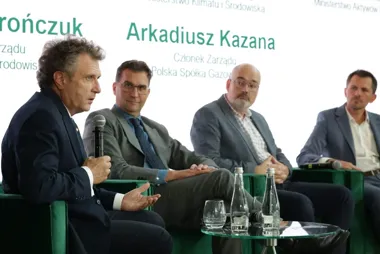 Czterech mężczyzn w garniturach siedzi na zielonych fotelach podczas panelu dyskusyjnego, jeden z nich przemawia do mikrofonu.