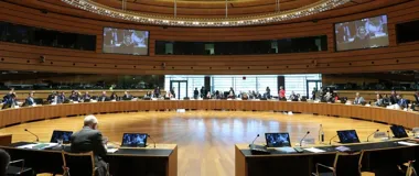 Posiedzenie w dużej, owalnej sali konferencyjnej z drewnianymi panelami, podczas którego uczestnicy siedzą przy zakrzywionym stole, a na ekranach wyświetlane są
