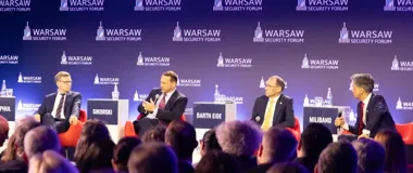 Czterech mężczyzn w garniturach uczestniczy w panelu dyskusyjnym podczas Warsaw Security Forum, siedząc na czerwonych fotelach przed publicznością.